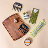 Flat lay of Gather & Graze gift box contents including a serving board, bear claw meat shredders, salami, hot honey, gourmet chocolate, and seasoning blend.