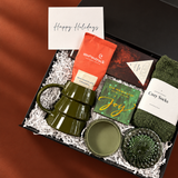 Holiday gift box with a Happy Holidays card, olive green tree mug, cozy socks, Moonstruck cocoa, Compartés chocolate, Joy matches, and green glass accessories.
