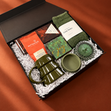 Modern holiday gift box filled with cozy green and cinnamon-toned gifts, including a ceramic tree mug, socks, Moonstruck cocoa, Compartés chocolate, matches, and glassware.