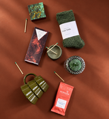 Holiday gift flat lay on a deep cinnamon background featuring cozy green socks, Moonstruck hot cocoa, Compartés chocolate, a Joy matchbox, olive ceramic tree mug, green glassware, and scattered matches.