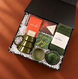 Angled view of a curated holiday gift box with an olive green mug, cozy socks, hot cocoa, chocolate, matches, and green glass pieces on a rich cinnamon background.