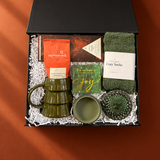 Open holiday gift box on a deep cinnamon background featuring an olive green tree mug, cozy green socks, Moonstruck hot cocoa, Compartés chocolate, a Joy matchbox, and green glass accessories.