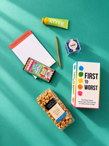 Flat lay of Game Night gift box contents including First to Worst, caramel popcorn, chocolate, candy, hand cream, Chaos Coordinator notepad and gold pen.