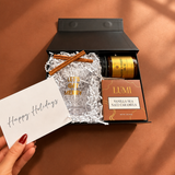 Small holiday cocktail gift featuring a Let’s Get Merry glass, hot buttered rum mix and vanilla sea salt caramels
